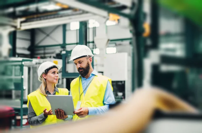 Two factory workers in vests and hard hats reviewing a clipboard and going over job costing information