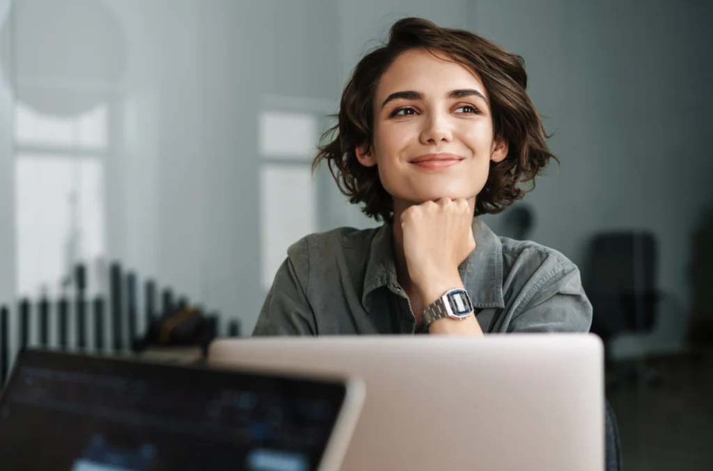 A woman looking slightly away from her laptop in a satisfied manner, visualizing the employee scheduling and retention benefits included