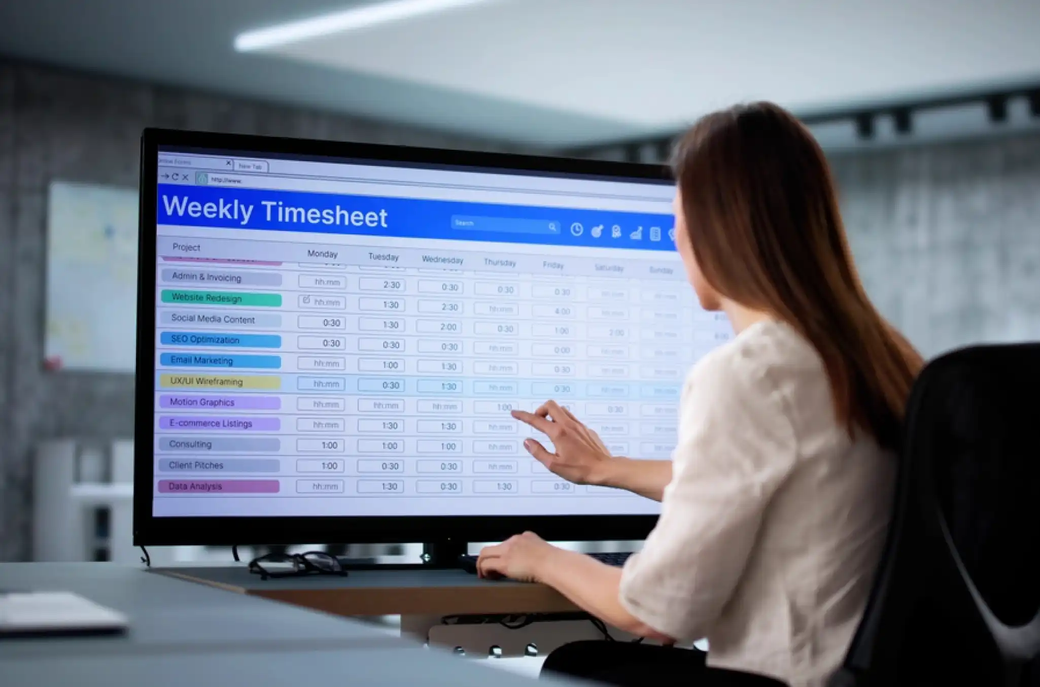A female worker looking at a wide screen computer monitor with the visual representation of how to calculate hours worked by going through a time tracking spreadsheet