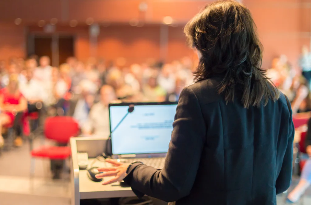 A teacher overlooking a classroom, camera is angled with her back to it, and there's a scheduling software that illustrates how to schedule employees in higher education because she's there and present