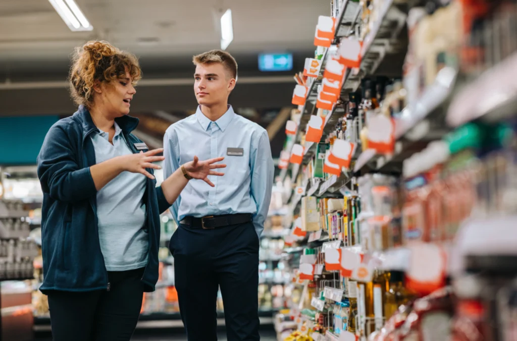A manager walking through a grocery store with a younger employee, talking about what time theft is and how it's important to clock hours properly