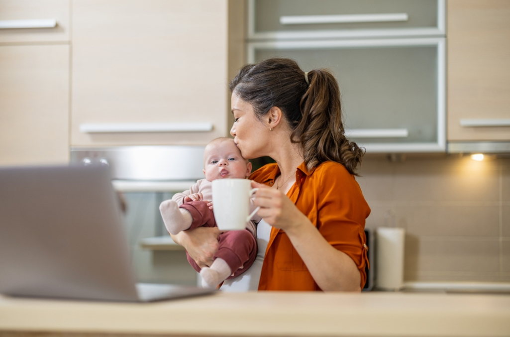 A woman at home, sitting at a kitchen island, holding her baby in one arm and a cup of coffee with the other hand, to represent time off through FMLA