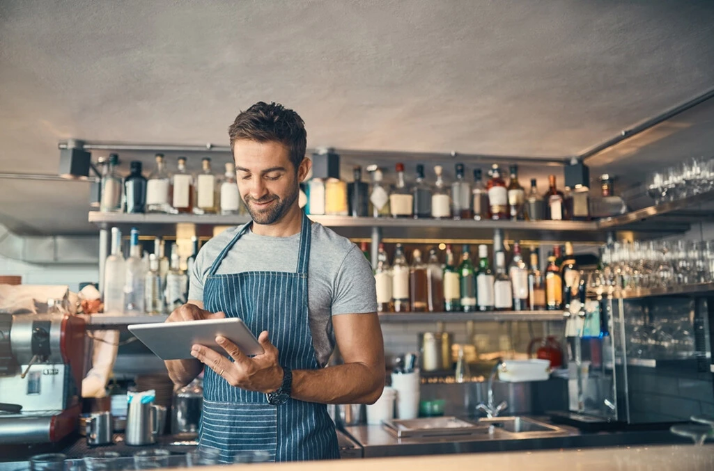A male bartender in a pinstripe apron reviewing inventory on a tablet and checking in on a schedule request made to be Fair Workweek Law compliant