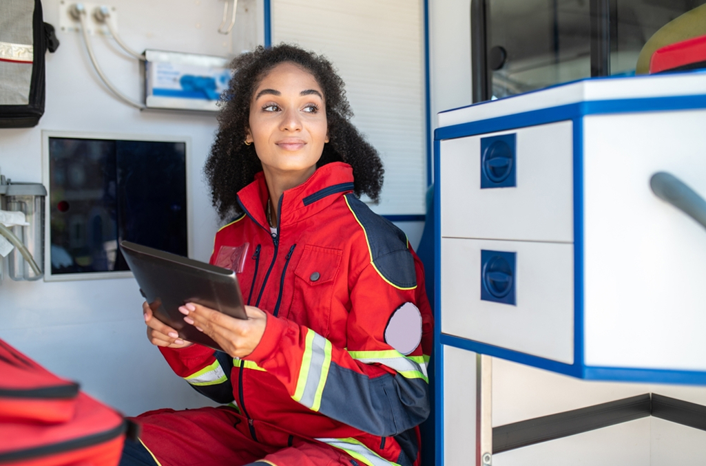 woman in red paramedic uniform sitting in back of an ambulance truck holding a tablet and looking into the distance
