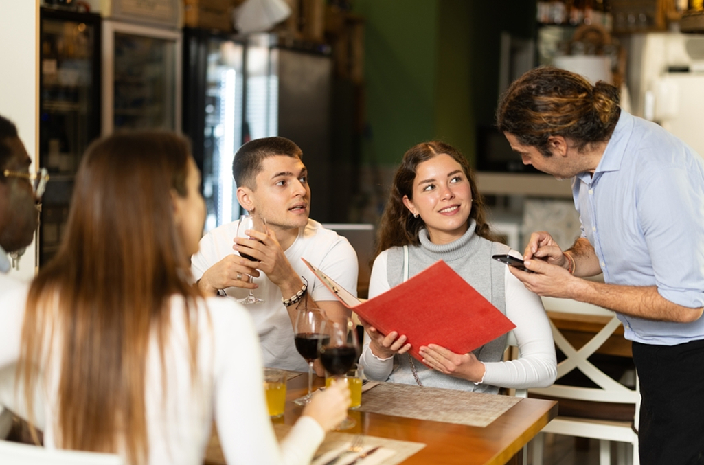 Three guests seated at a table in a restaurant, one holding a red menu, server is leaning over to take order