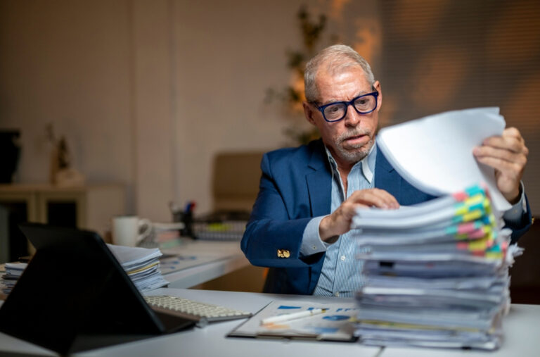 A payroll professional looking at a stack of paperwork at a desk, which is full of timesheet errors, as he sits with a concerned and confused demeanor