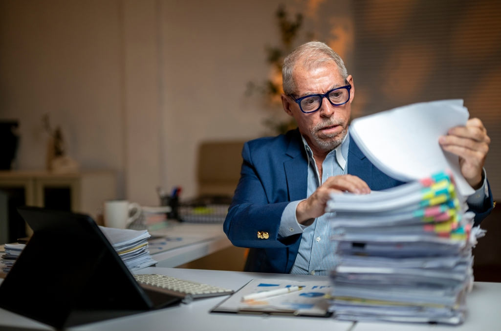 A payroll professional looking at a stack of paperwork at a desk, which is full of timesheet errors, as he sits with a concerned and confused demeanor