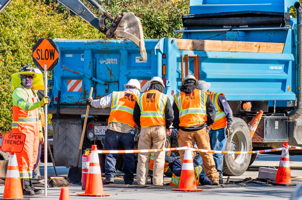 City public workers wearing reflective orange and yellow vests looking into the street with bright blue dump truck behind them