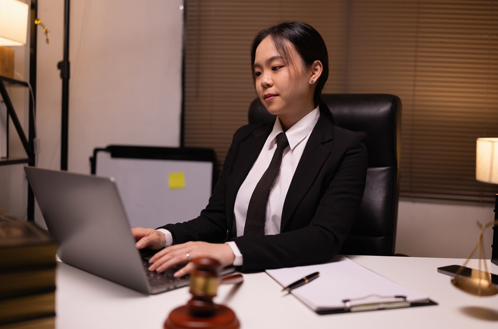 Women sitting at a desk with suit and tie on, typing on an open laptop with gavel and justice scales on the desk