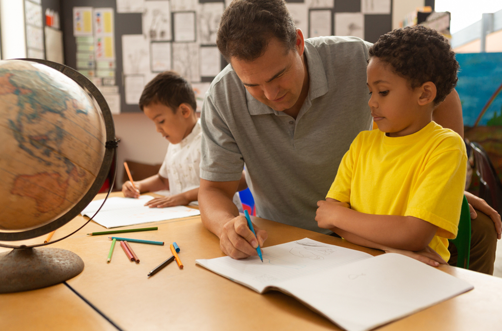 Male teacher holding a marker over an open book on a wooden desk while elementary age child in bright yellow shirt looks on. Desk is covered with colored pencils and a globe in view