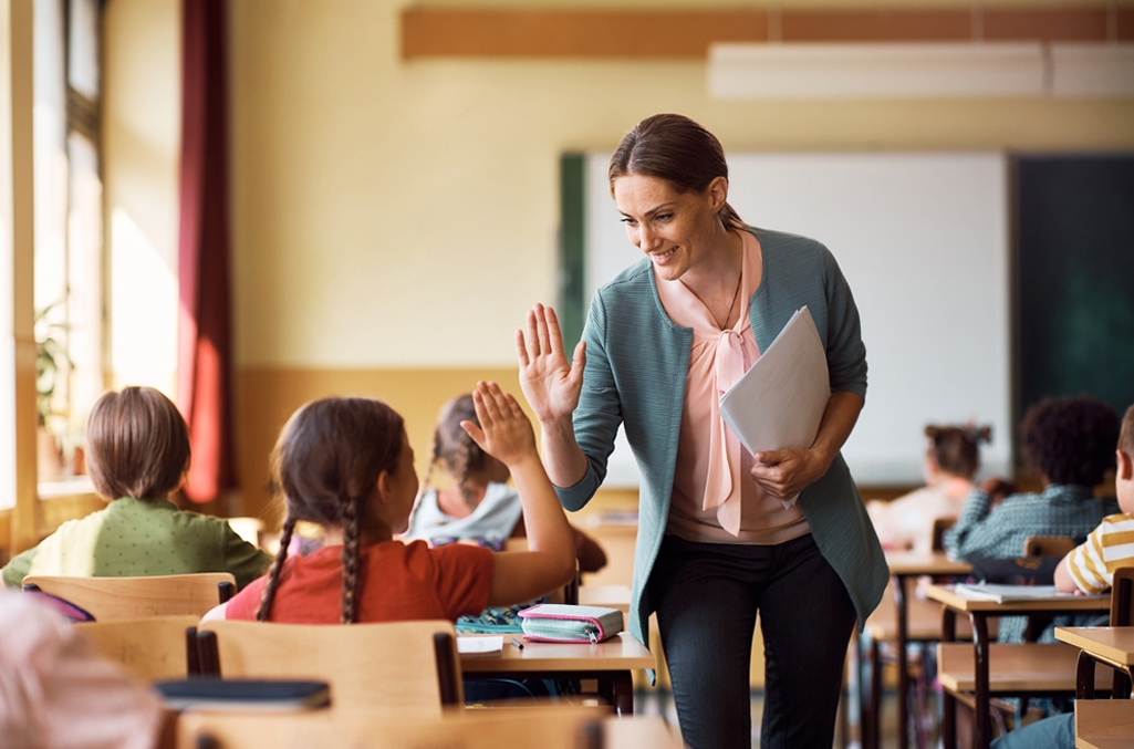 Female teacher standing in a classroom wearing light blue sweater and light pink button down shirt high-fiving an elementary age girl student with pigtails seated at her desk