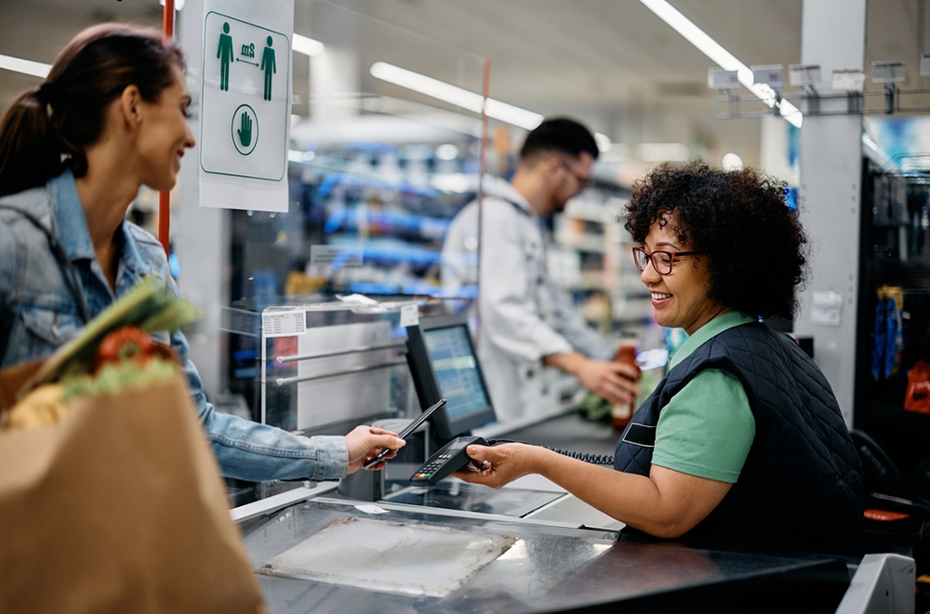 Cashier at a grocery store on one side of the conveyor belt handing credit card to female customer