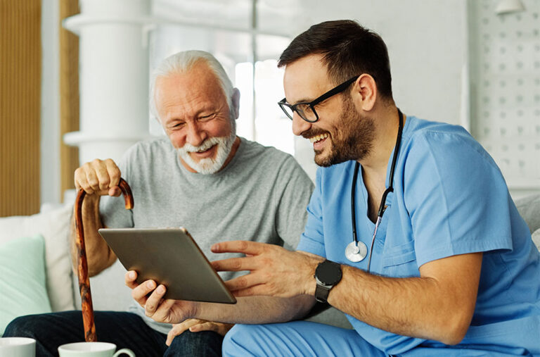 Doctor or nurse caregiver in light blue scrubs showing a tablet screen to senior man and laughing at home or nursing home