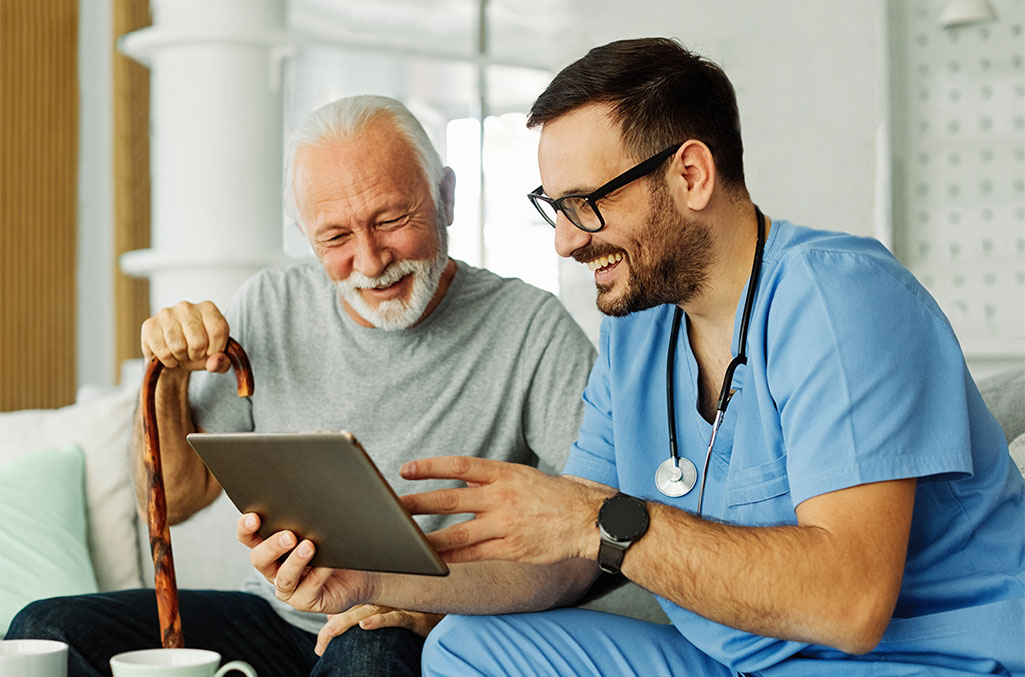 Doctor or nurse caregiver in light blue scrubs showing a tablet screen to senior man and laughing at home or nursing home