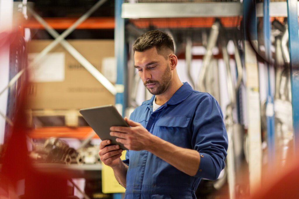 Man in blue jumpsuit at a distribution facility or mechanic shop looking at a tablet