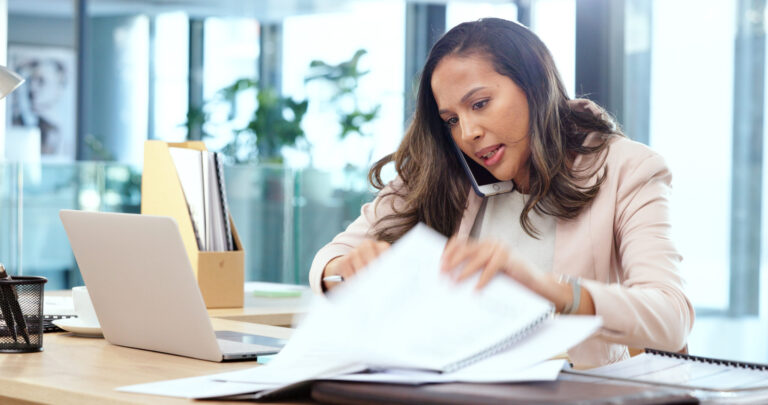 Female advertising agent with dark hair looking busy while talking on a phone call and searching through documents on a messy table in an office. Female marketing professional trying to find an important form.