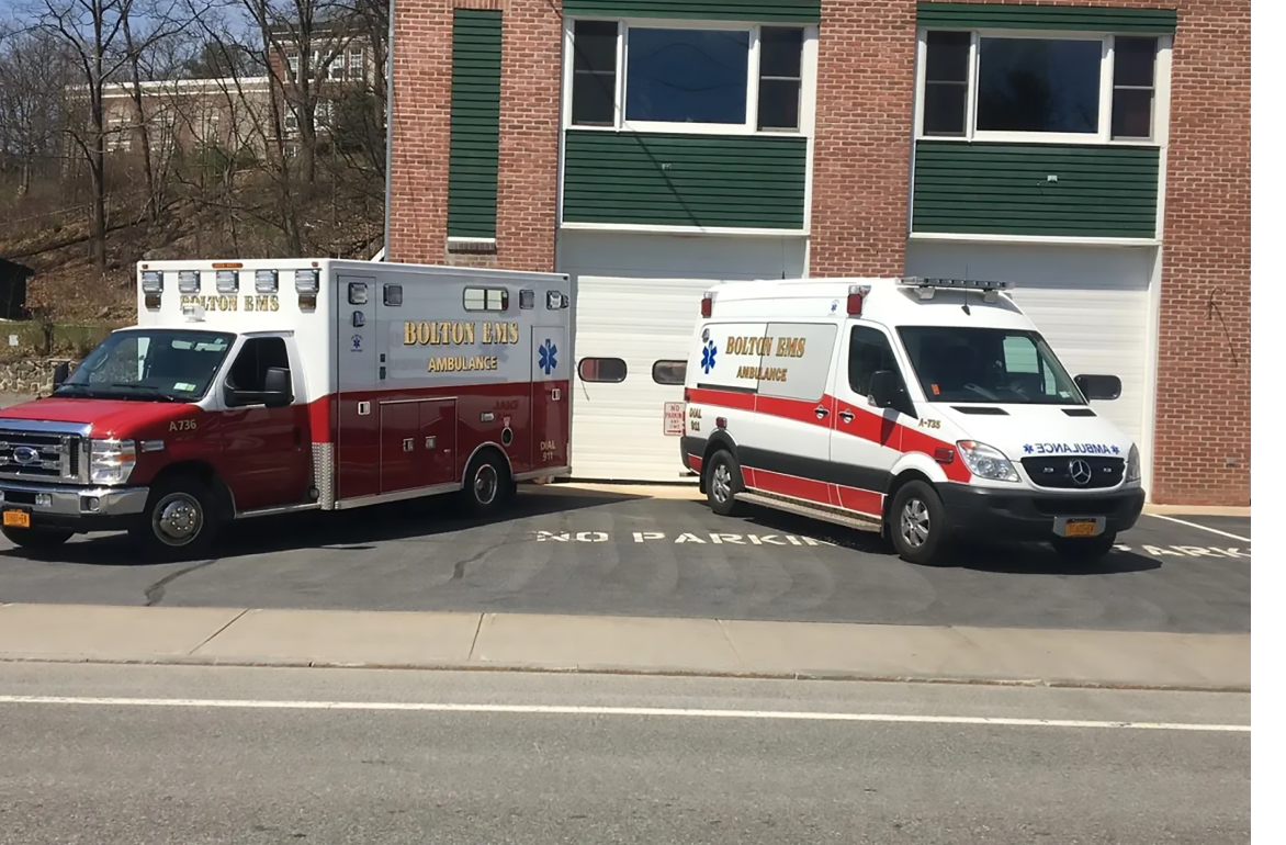 Two EMS vehicles parked with nose of vehicles pointed to the left and right hand corner to make a V shape and parked in front of a closed garage at an EMS station