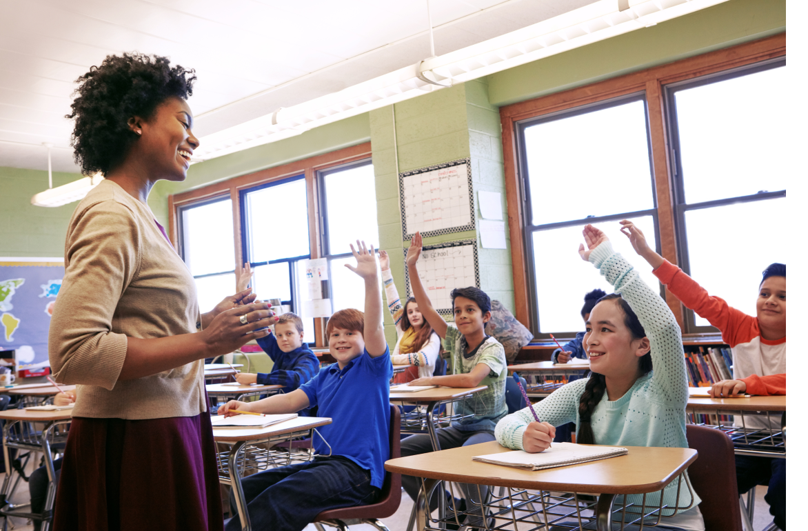 Teacher with short dark hair on the left side standing in front of a classroom of elementary school children with hands raised