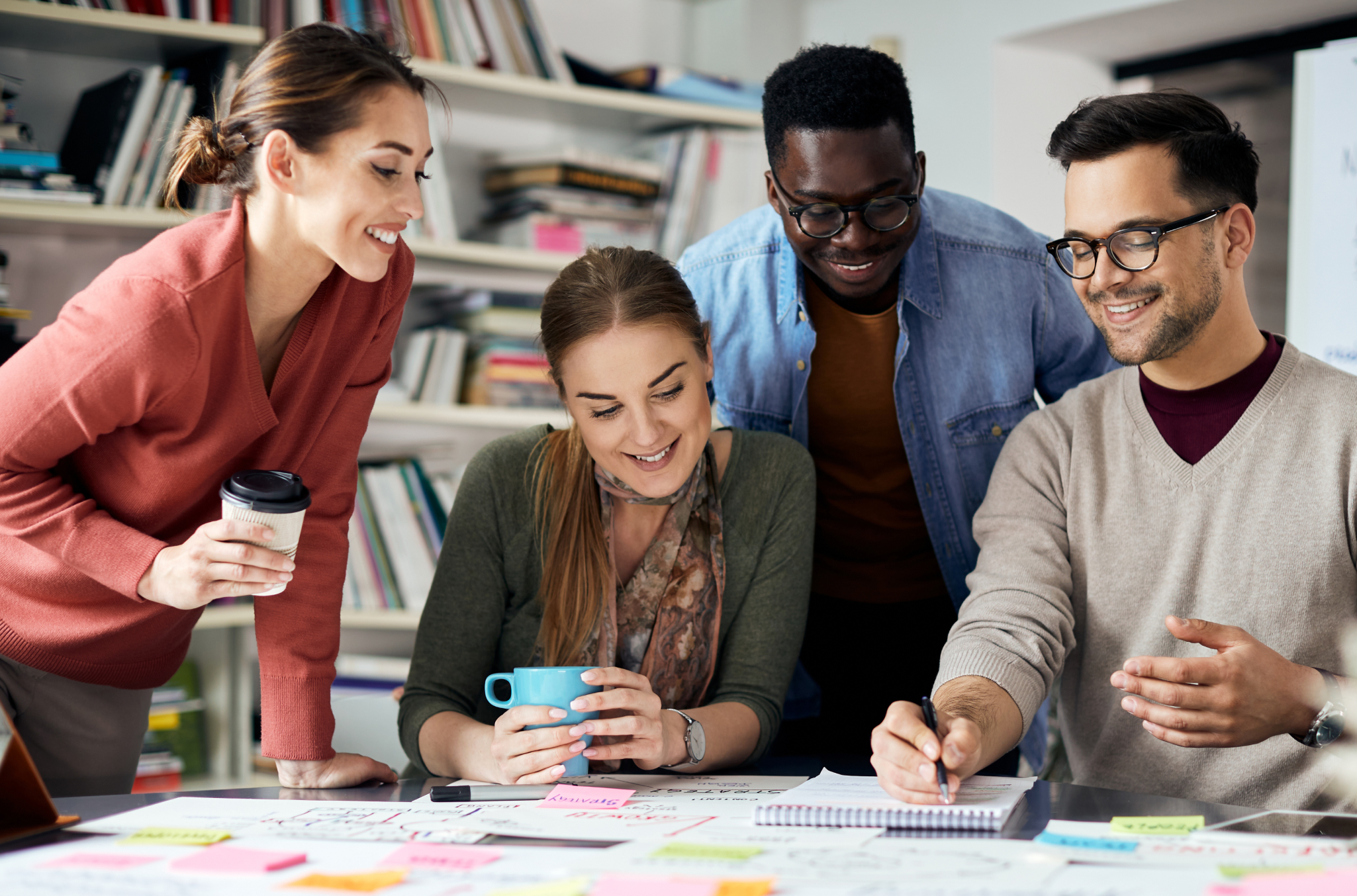 Four coworkers seated at a desk looking at a large weekly schedule with colored sticky notes on it